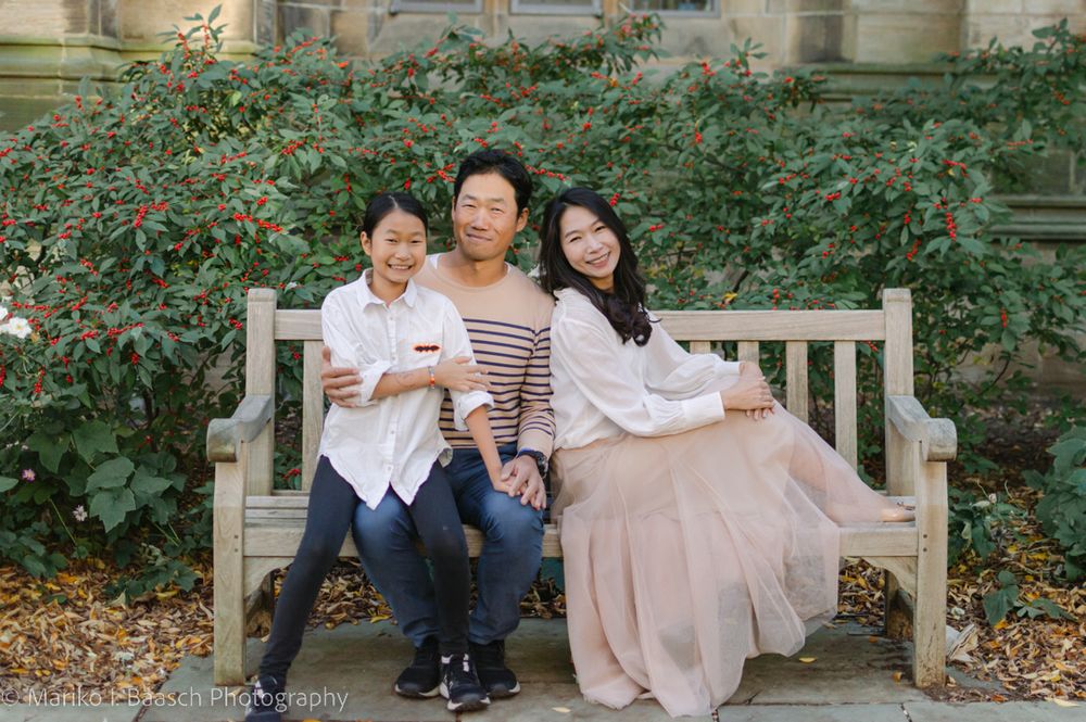 A family of three sits together on a stone bench surrounded by lush greenery.