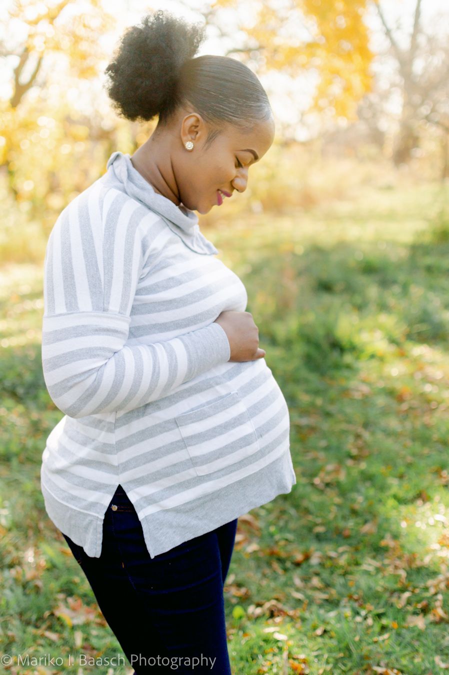 Pregnant Black woman in striped sweater cradling her baby bump outdoors in autumn.