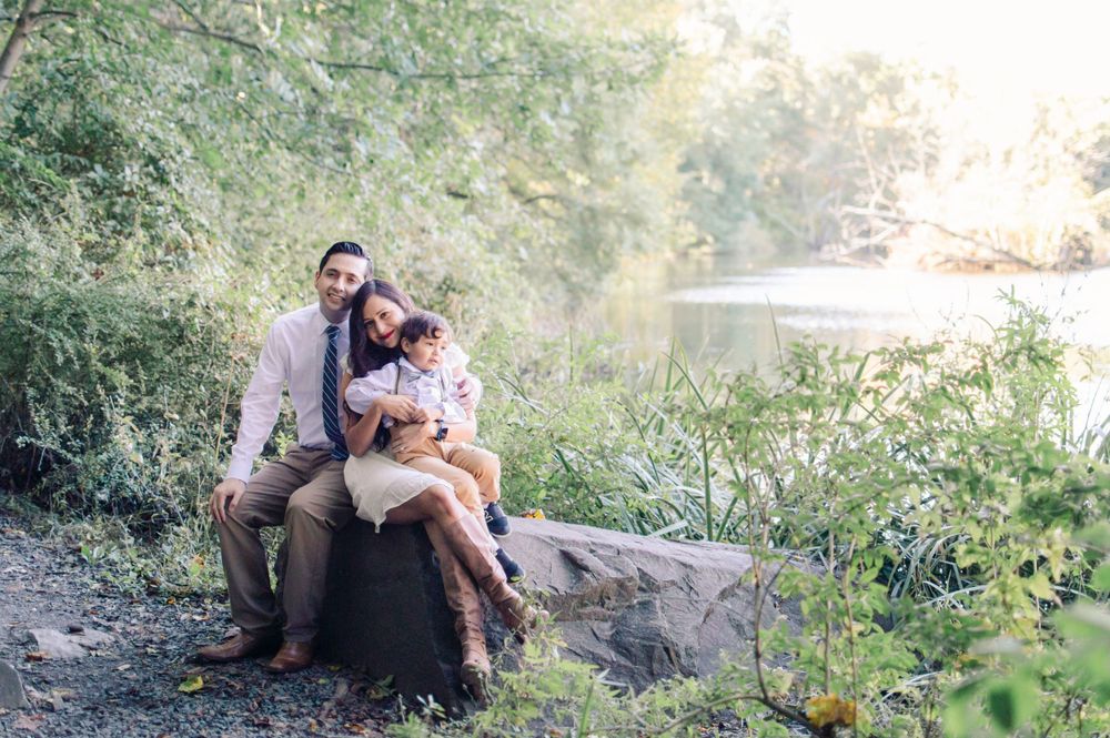 A happy family of three sits together on rocks beside a serene river surrounded by greenery.