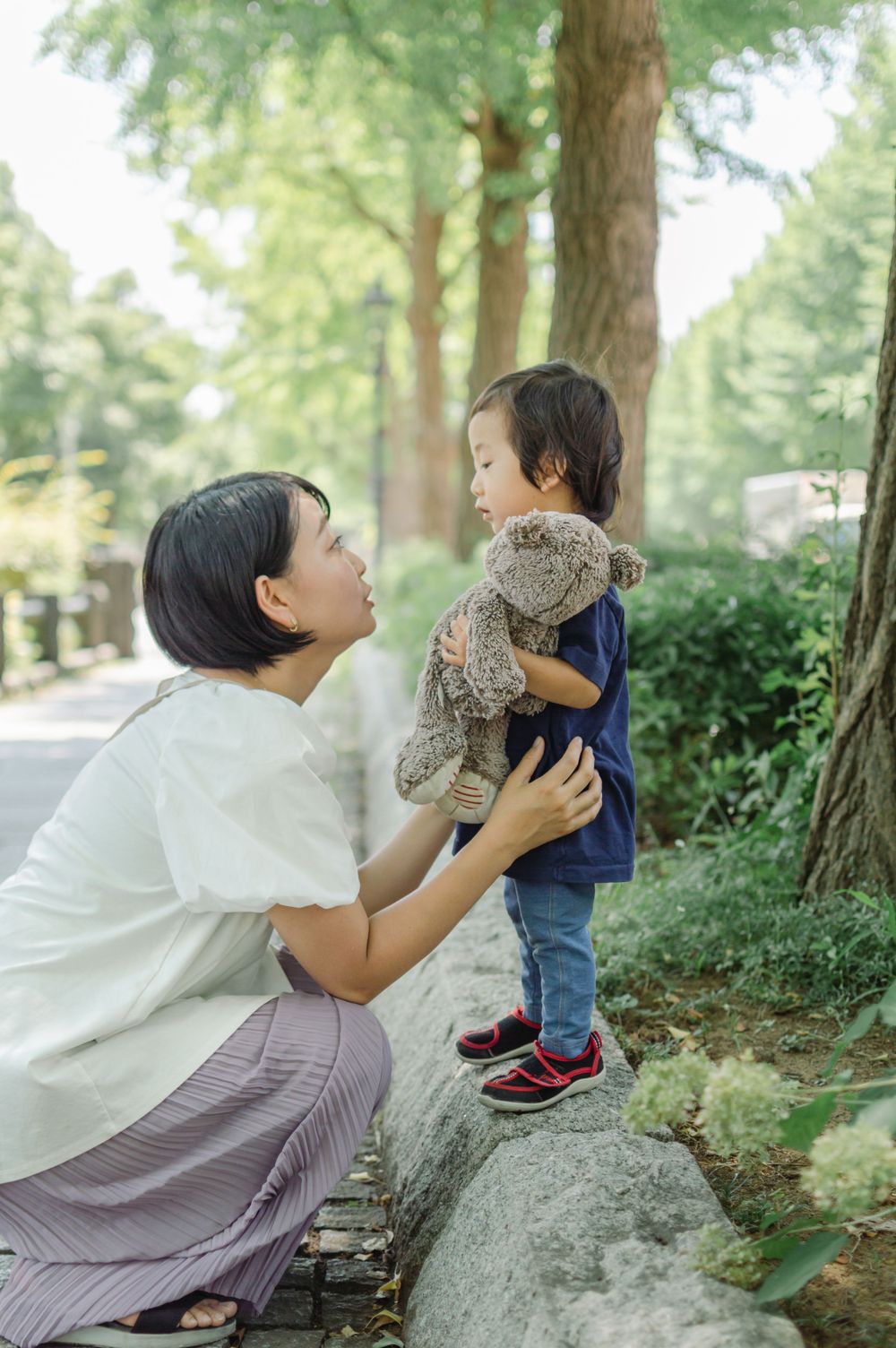 Asian mother crouching to talk with her young child holding a teddy bear in a park.