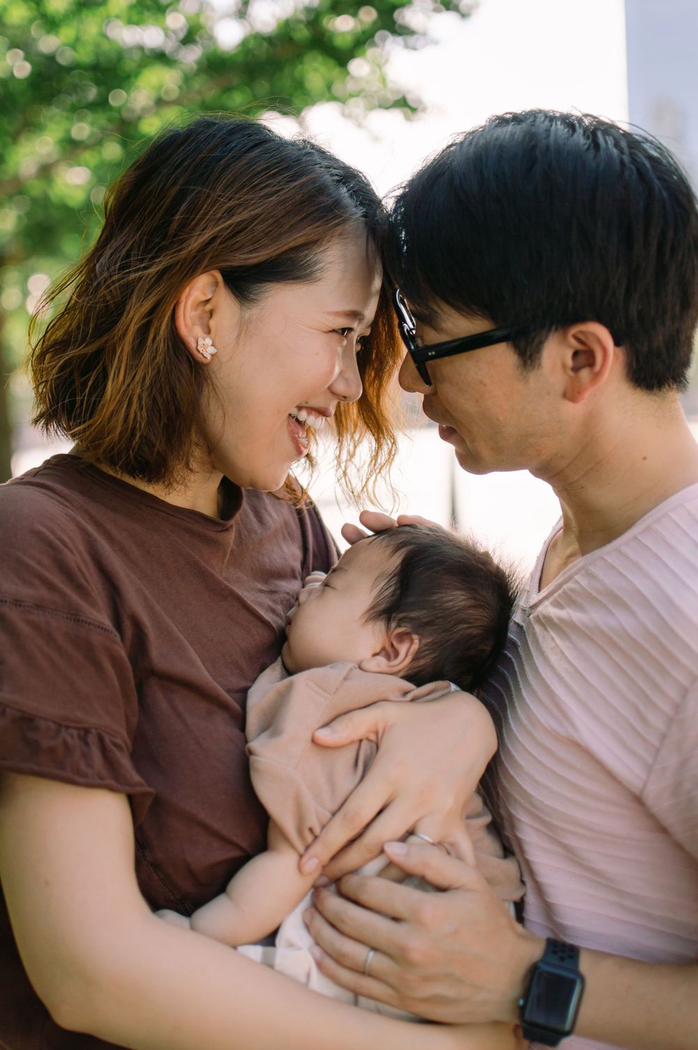 Smiling Asian couple touching foreheads tenderly while holding their sleeping newborn baby outdoors.