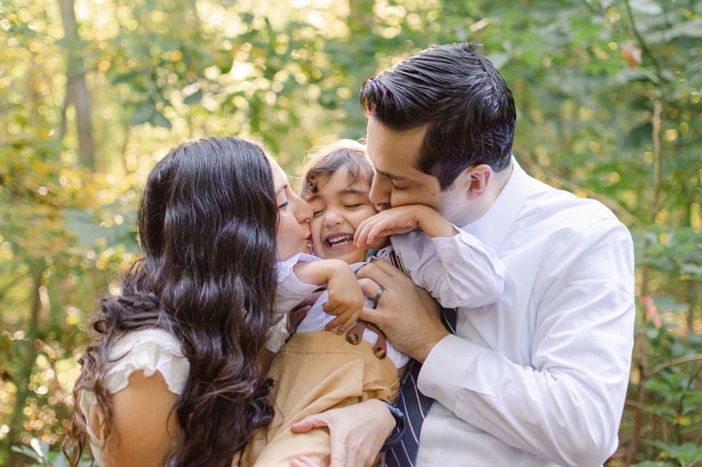 A joyful family moment as parents kiss their laughing toddler in a lush green forest setting.
