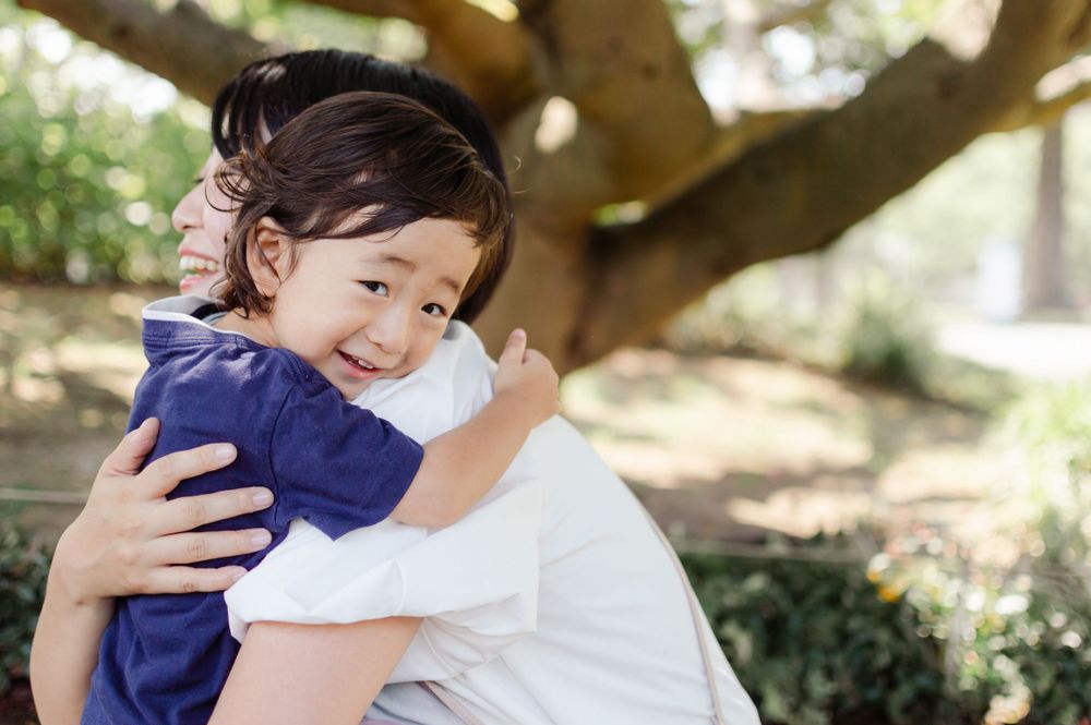 Happy young Asian child smiling while being hugged outdoors under a large tree.