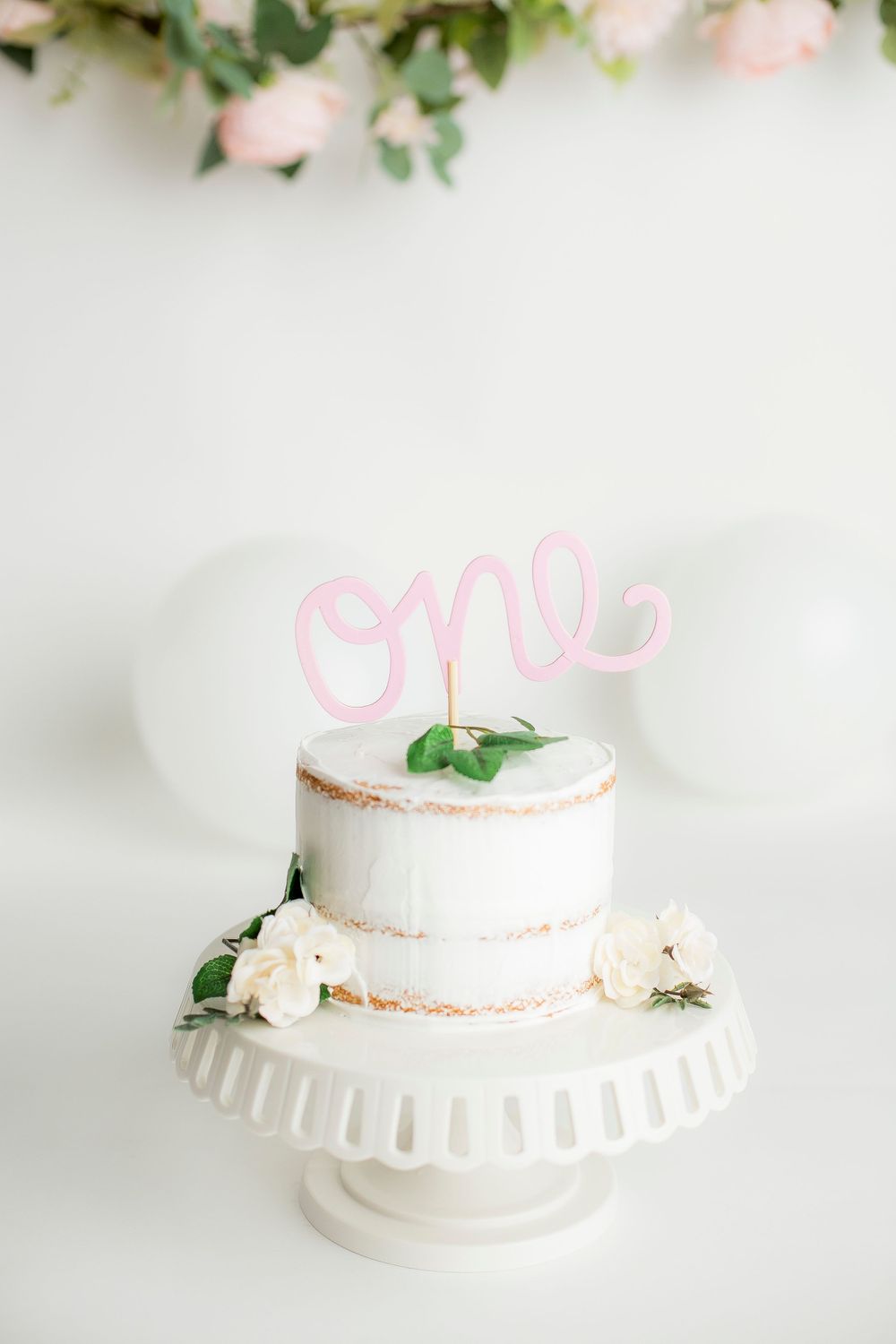 Simple white cake with pink one on top.  Cake stand is white and there is a pink garland hanging behind.