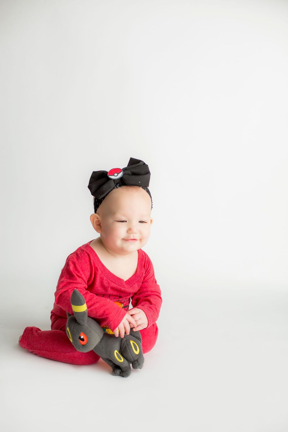 One year old girl sitting on white backdrop in a pokemon onesie and umbreon stuffy.