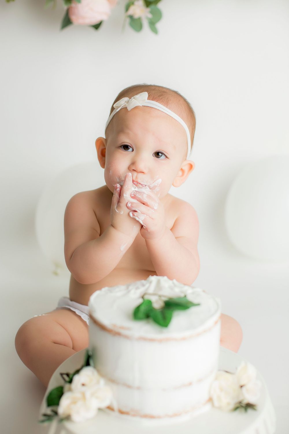 One year old baby girl sitting on white backdrop with her cake in front of her licking the cake off her fingers.
