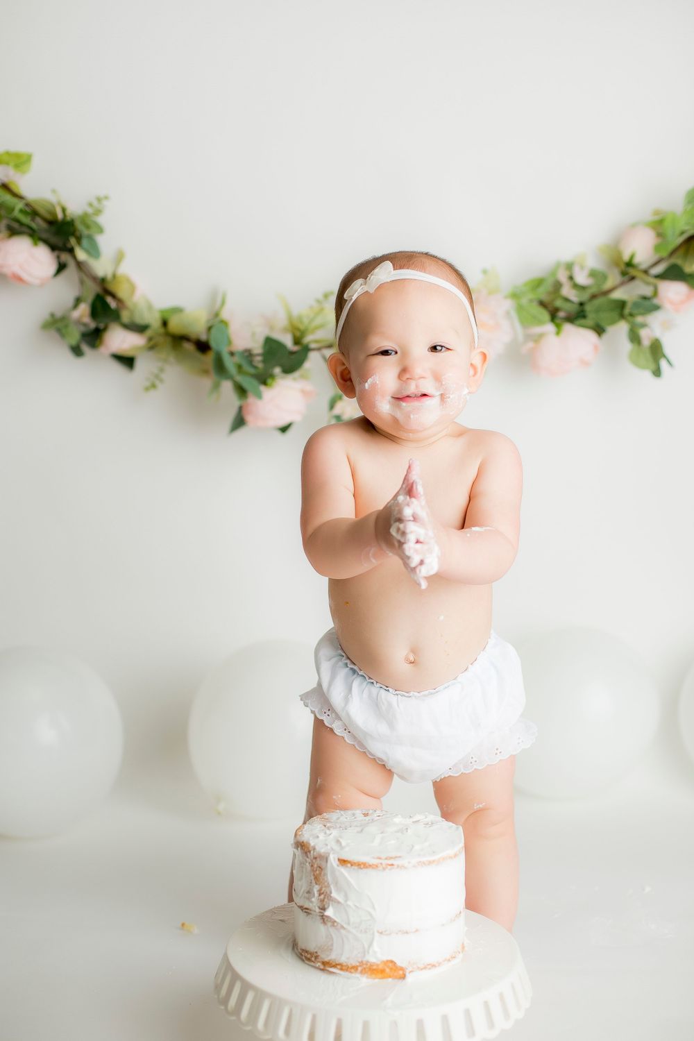 One year old baby girl standing above her white cake smiling and clapping.