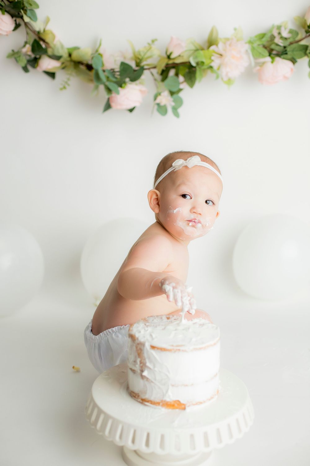 One year old baby girl in white bloomers squatting with white balloons behind her and white cake in front of her.