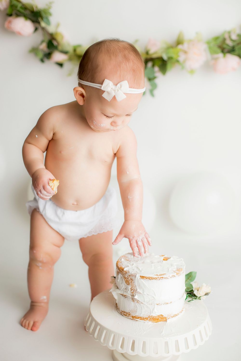 One year old baby girl standing in white bloomers looking and touching her birthday cake.