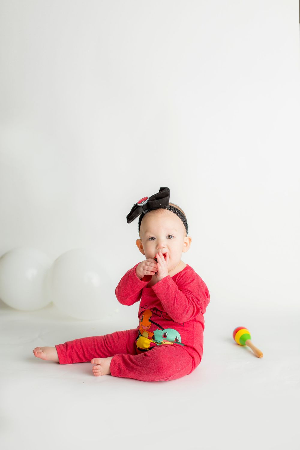 One year baby girl sitting on a white backdrop in a pokemon onesie with white balloons behind her.