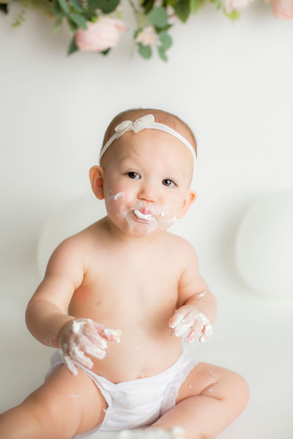 One year old baby girl sitting on white backdrop in white bloomers with cake on her face and hands.