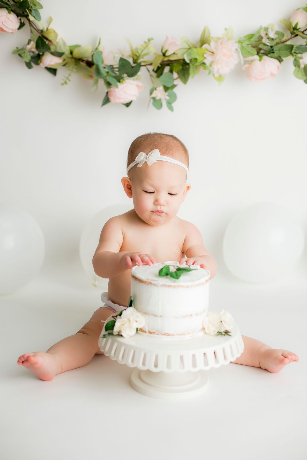 One year old baby girl on white backdrop with balloons and floral garland in background and cake in front of her.