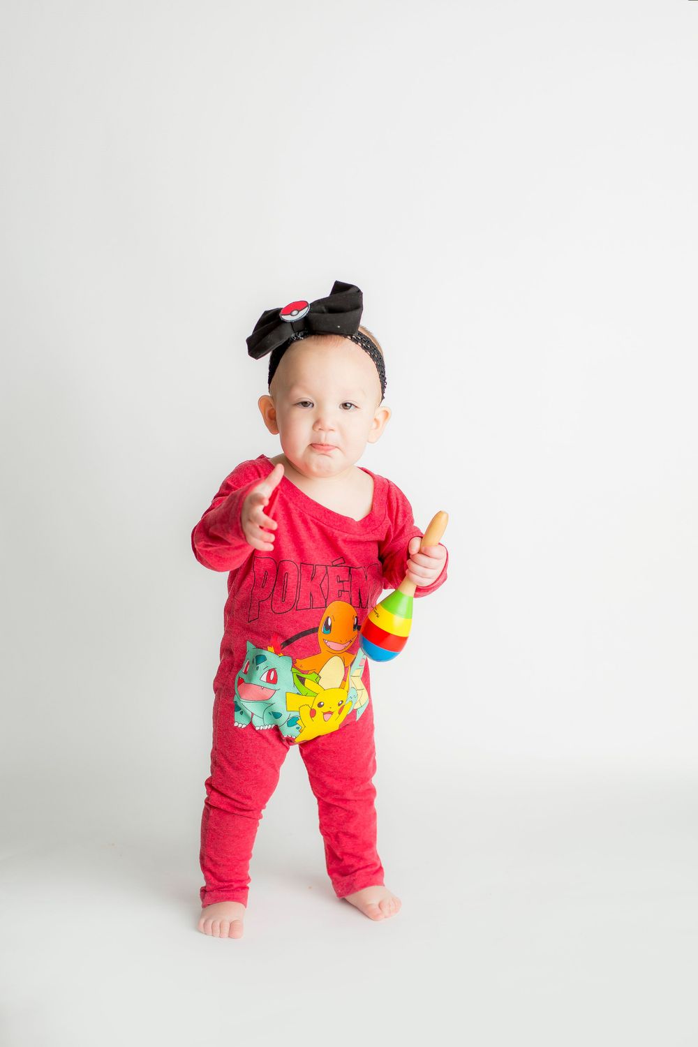 One year old baby girl standing on a white backdrop with a pokemon onesie and black bow headband.