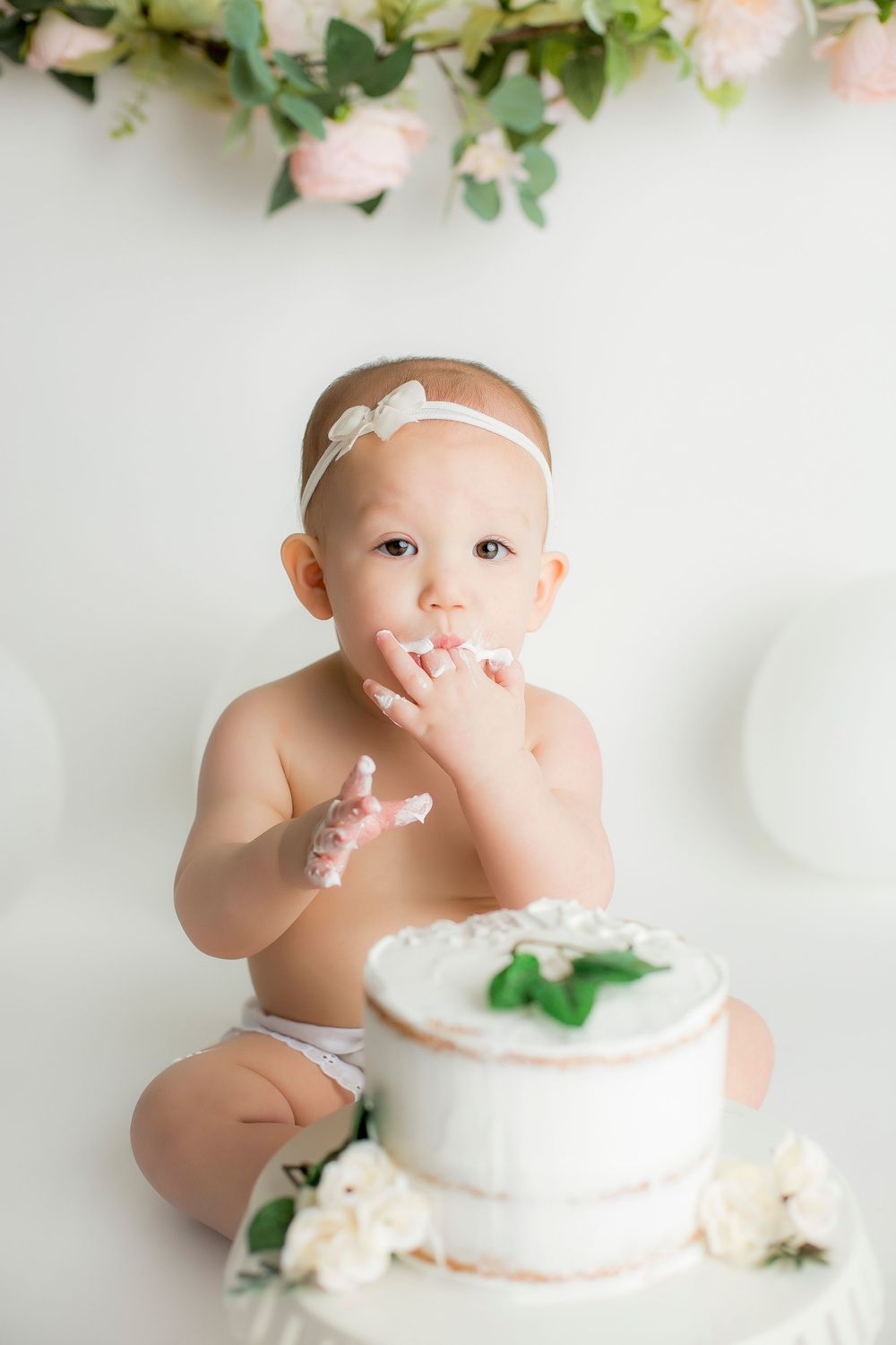 One year old baby girl sitting on white backdrop looking at camera licking cake and icing off her fingers.