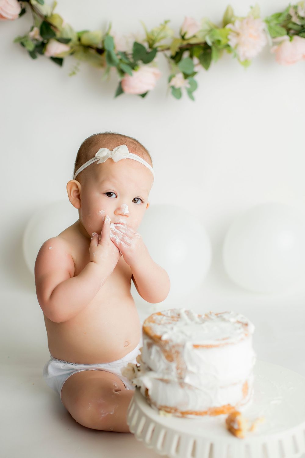 One year old girl sitting on white backdrop in white bloomers enjoying white birthday cake.
