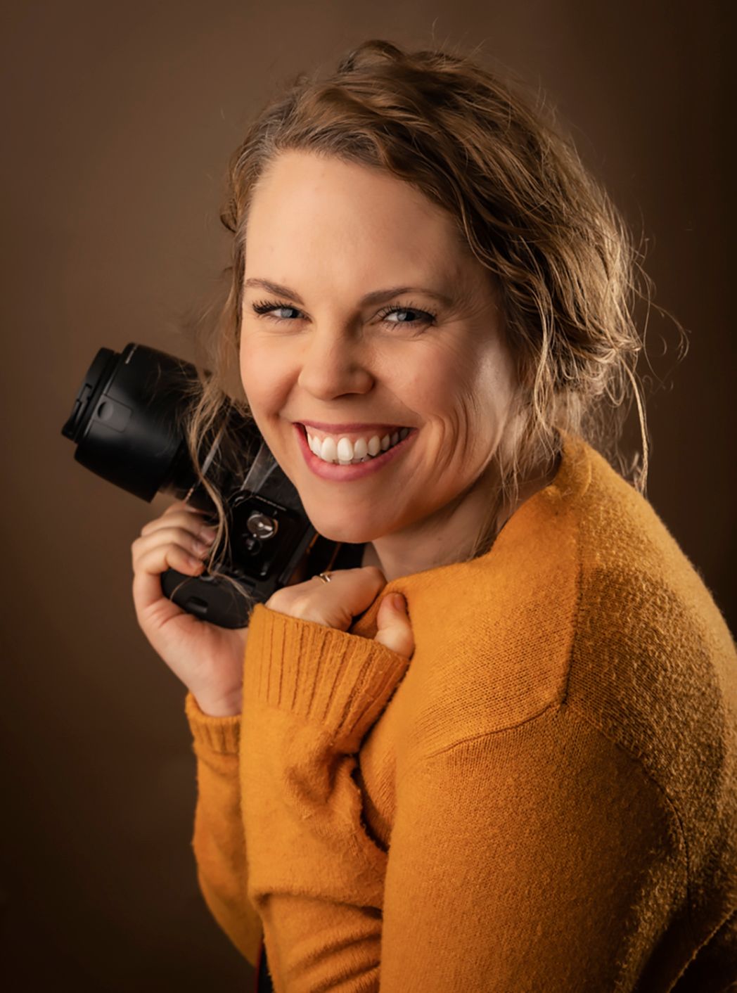 Portrait of photographer in orange sweater holding camera and smiling warmly against brown background.