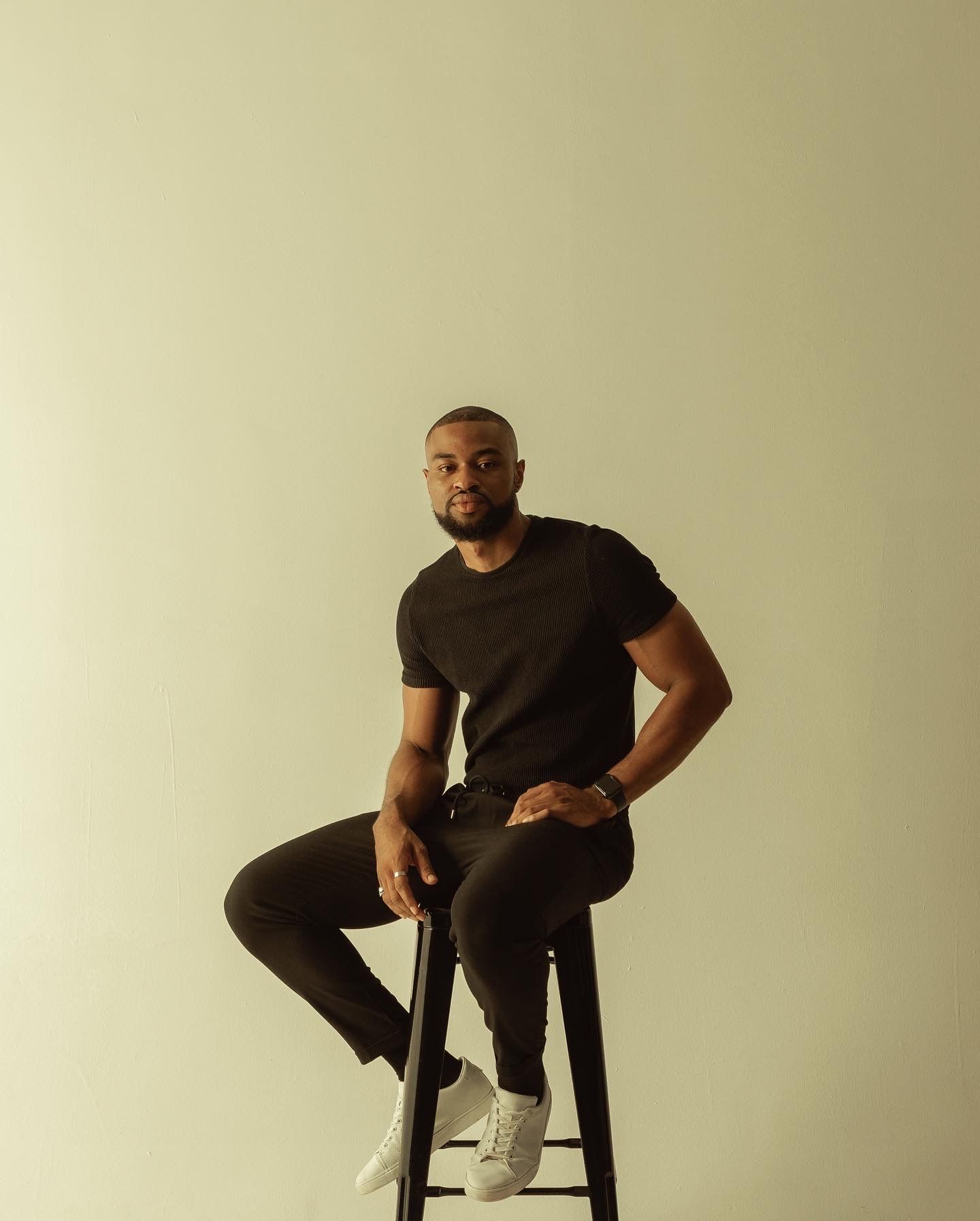 Minimalist portrait of a person seated on a stool against a beige background wearing dark clothing.
