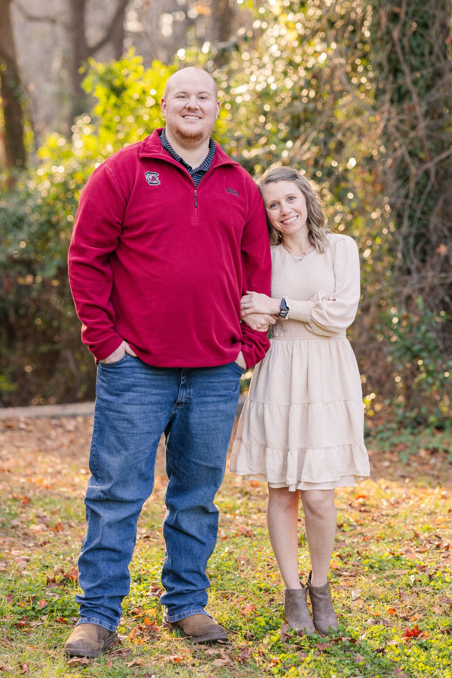 A couple poses together outdoors in fall, wearing red and beige against an autumn background.