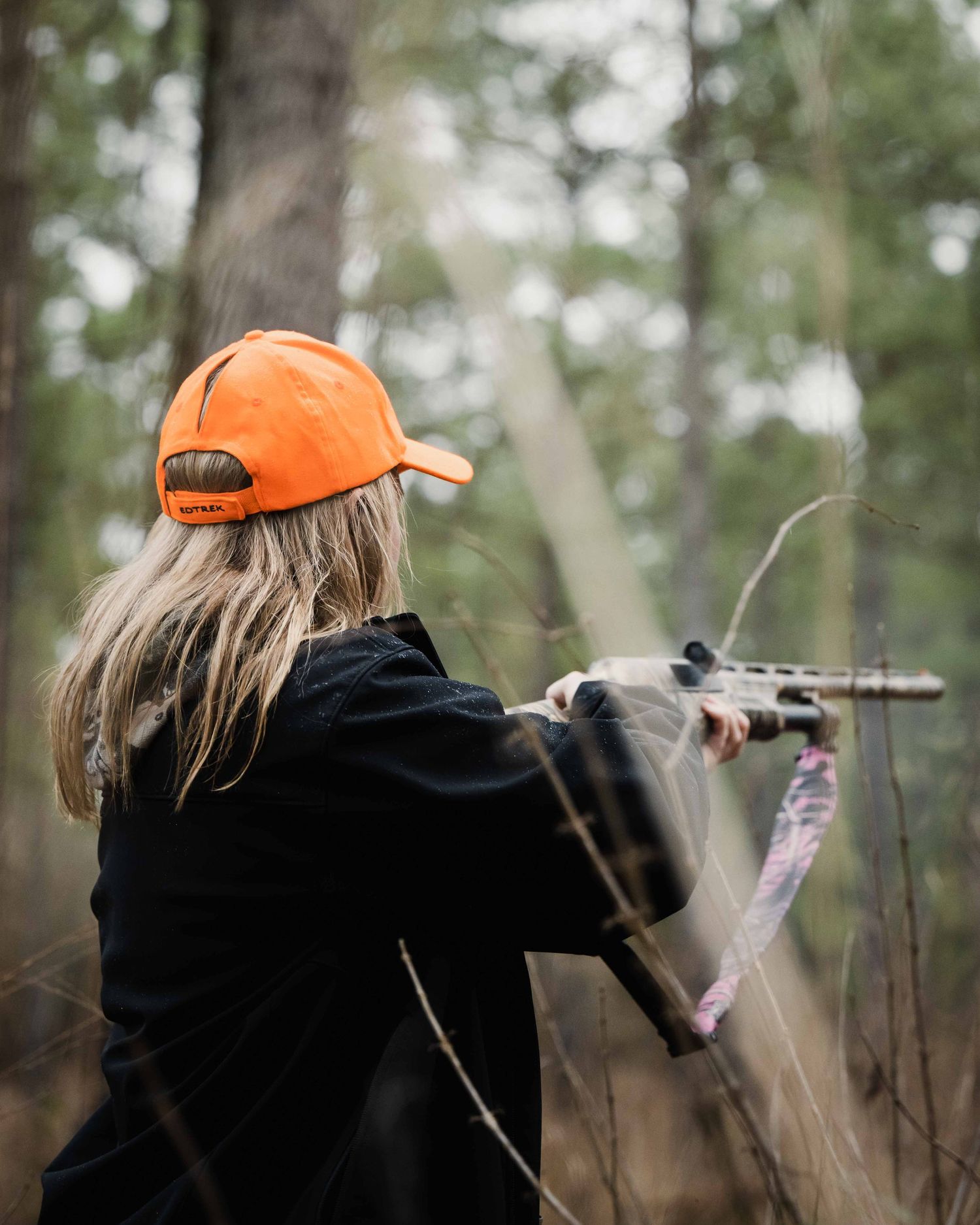 Hunter in orange safety cap aiming rifle in wooded area.