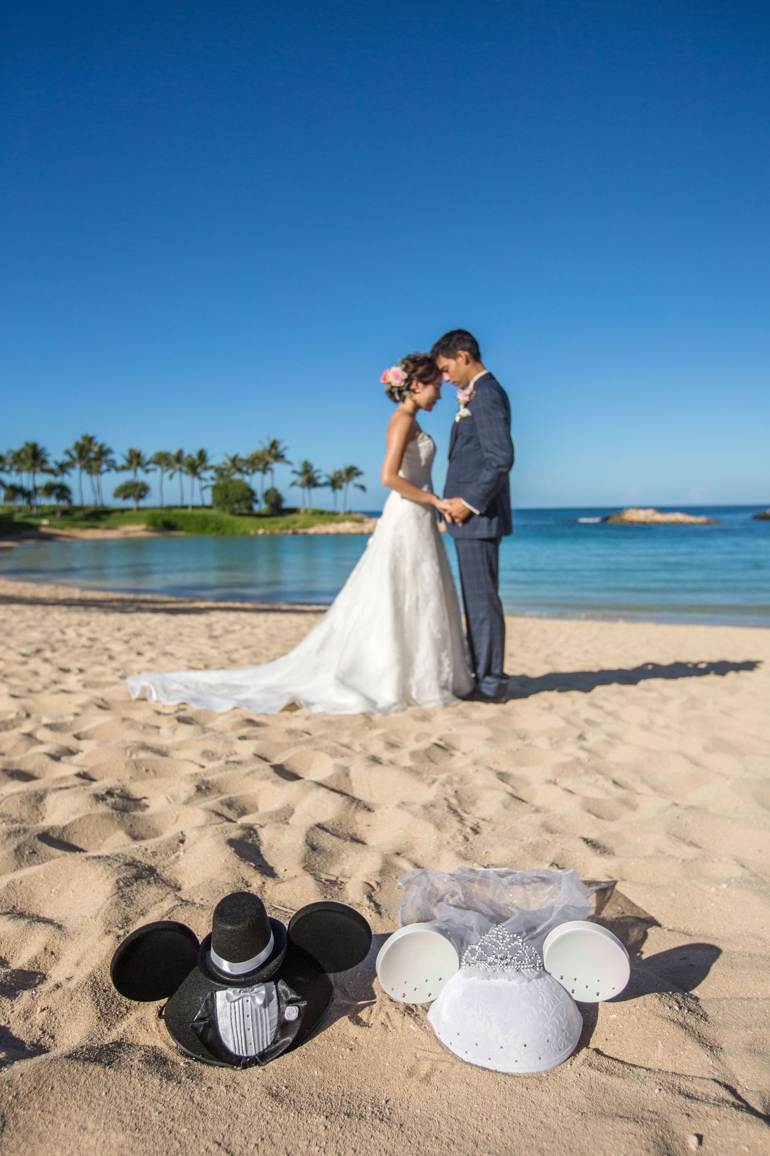 Bride and groom embracing on a sandy beach with Mickey Mouse ear hats in the foreground.
ビーチウェディングフォト
ハワイウェディング
ハワイ挙式
