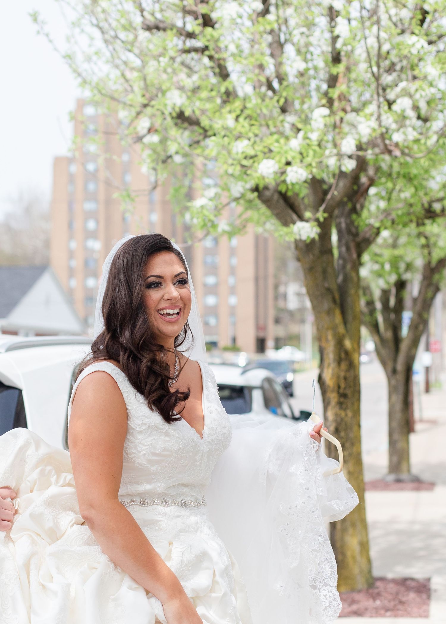 Bride in white dress posing joyfully outdoors near blooming spring trees.