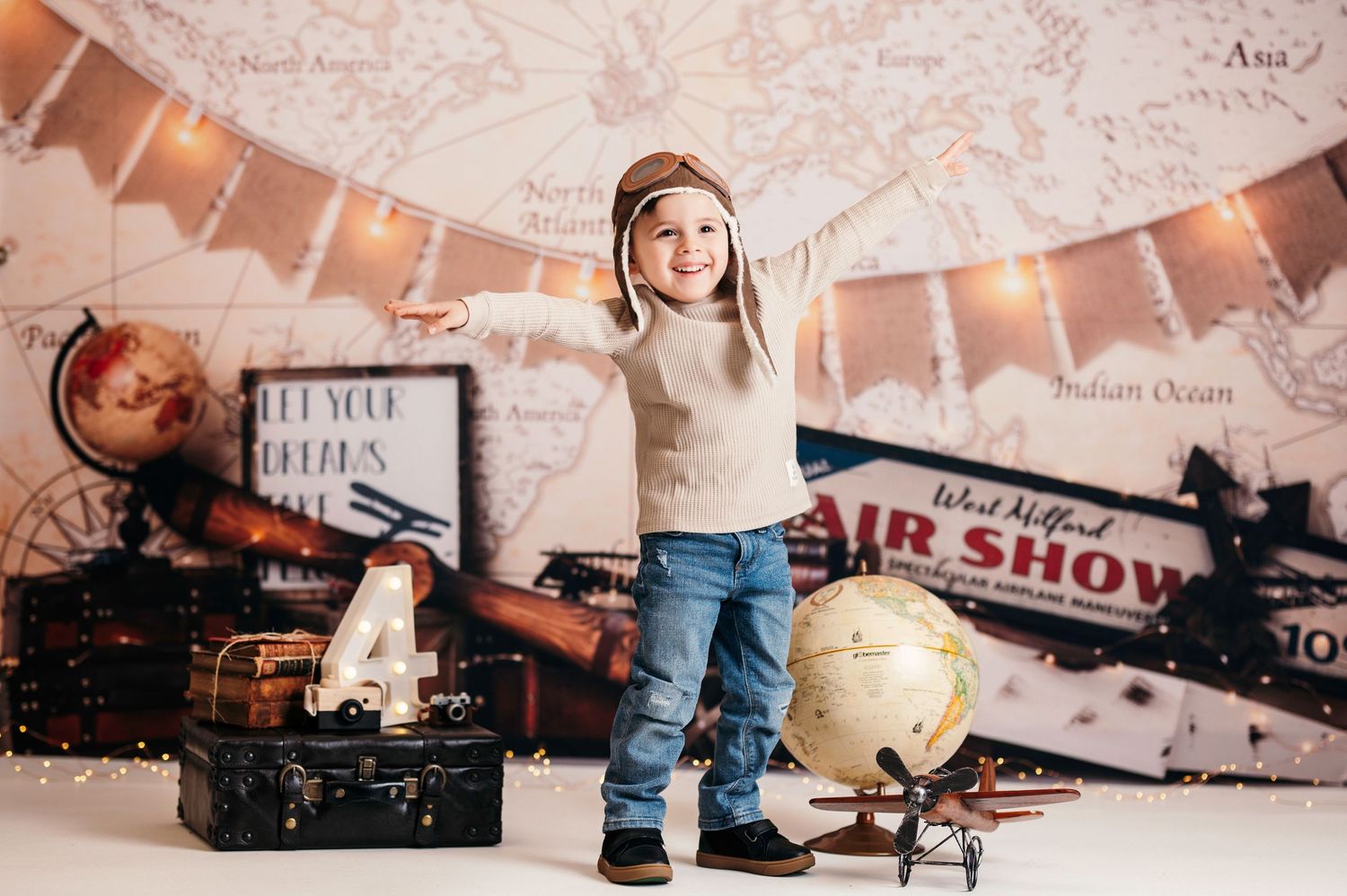 A joyful child in a pilot hat poses with a globe and vintage suitcase against an adventure-themed backdrop.