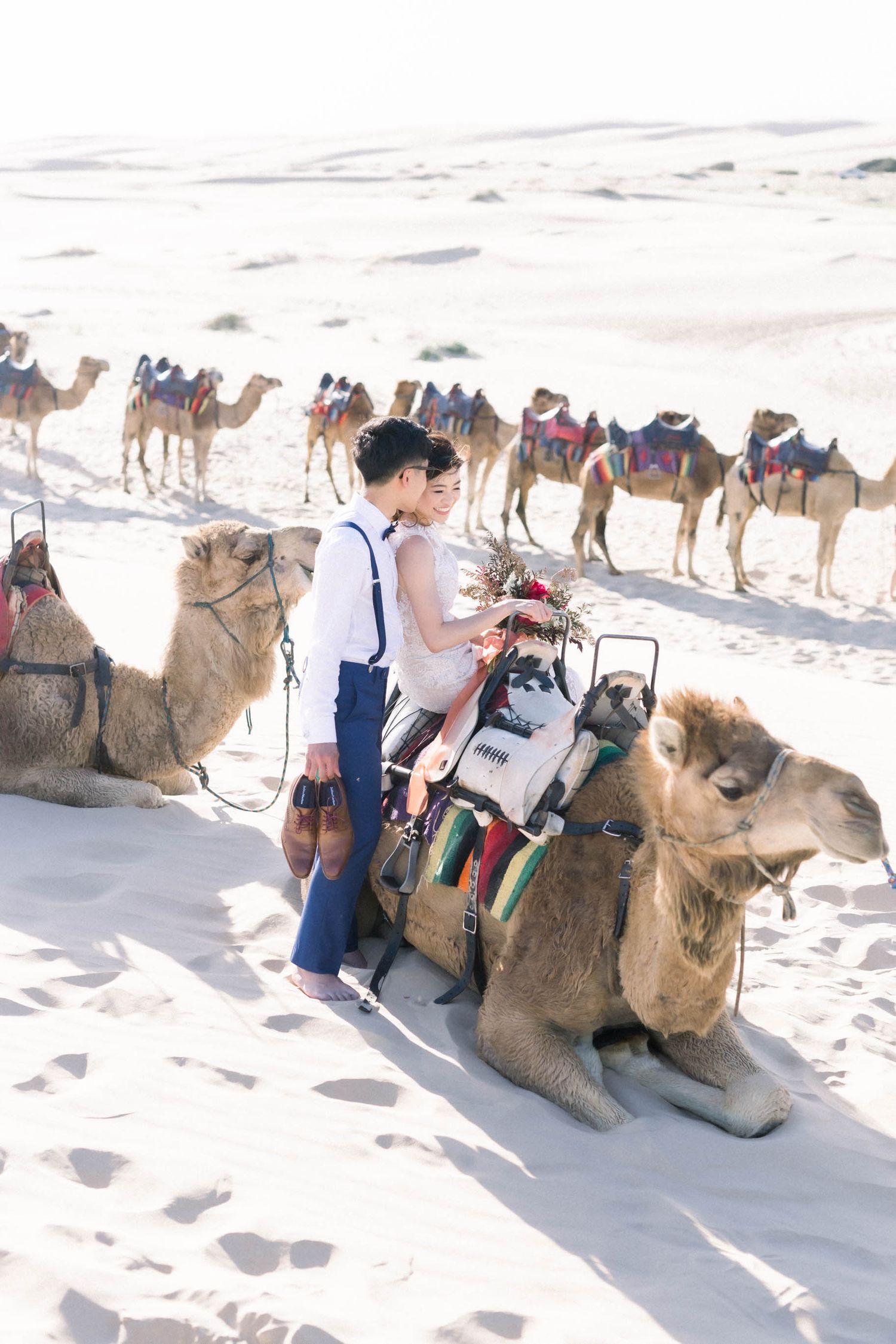 A unique desert wedding photo featuring a camel caravan and couple against white sand dunes.