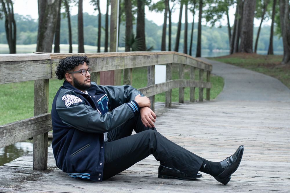 Graduate in casual attire sits on wooden boardwalk overlooking wooded area.