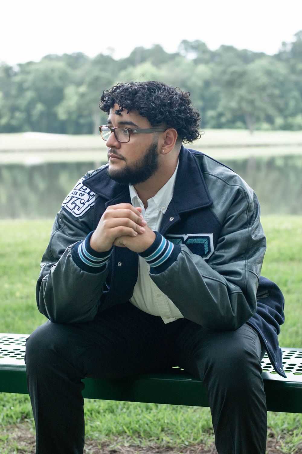 Graduate in varsity jacket sits thoughtfully on park bench near lake.