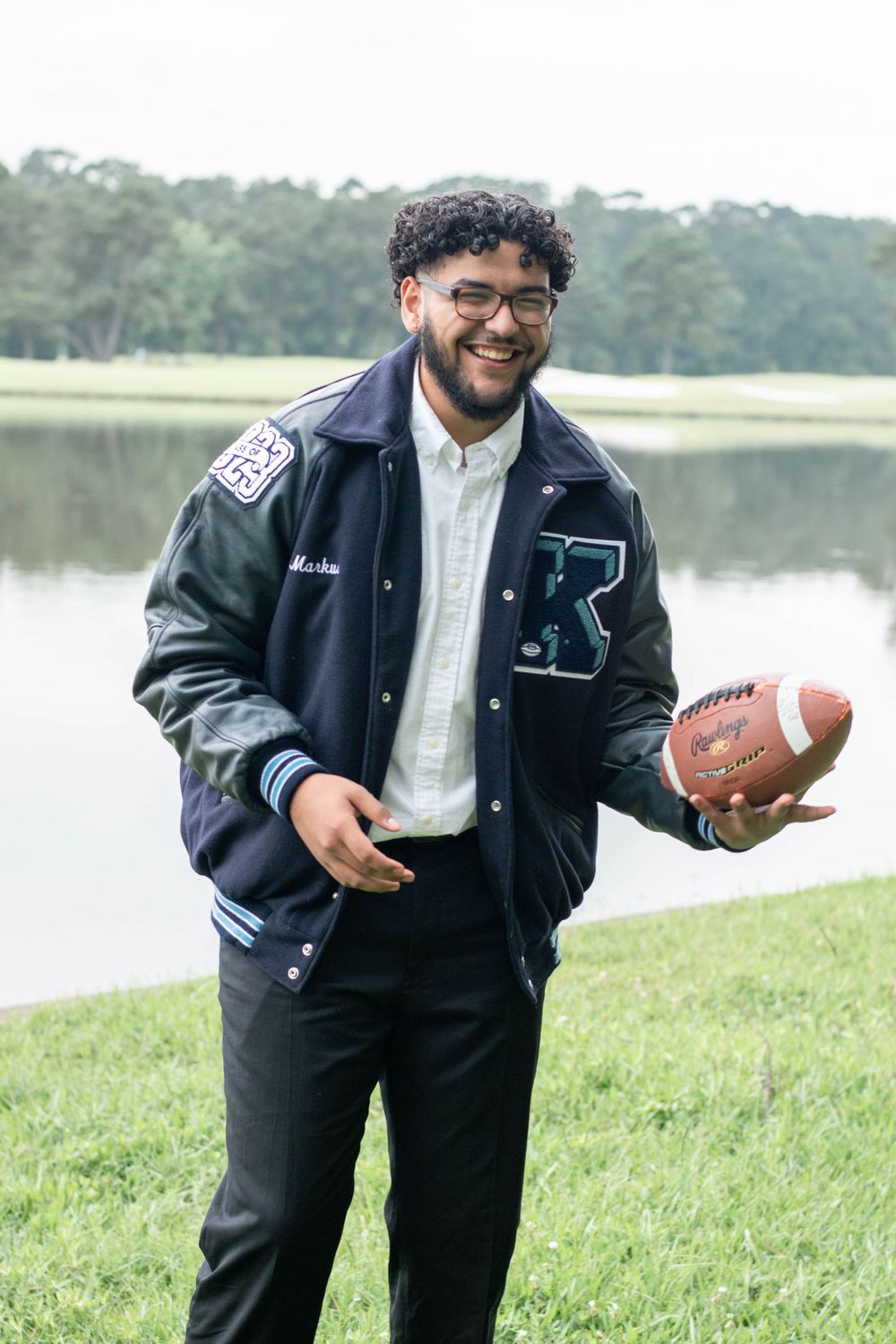 Graduate in varsity jacket smiles while holding football near scenic lake.