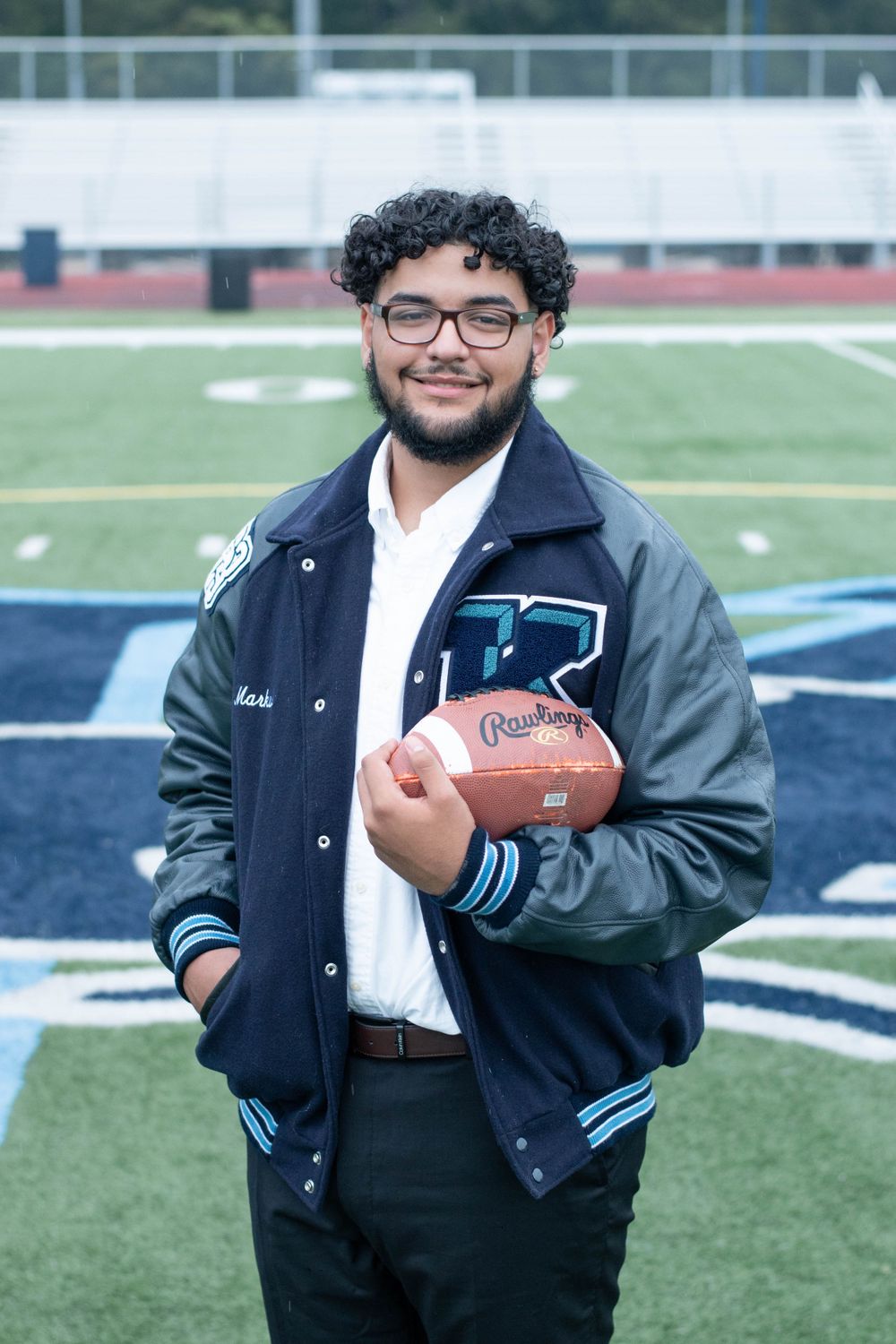 Graduate poses with football on field wearing varsity jacket.