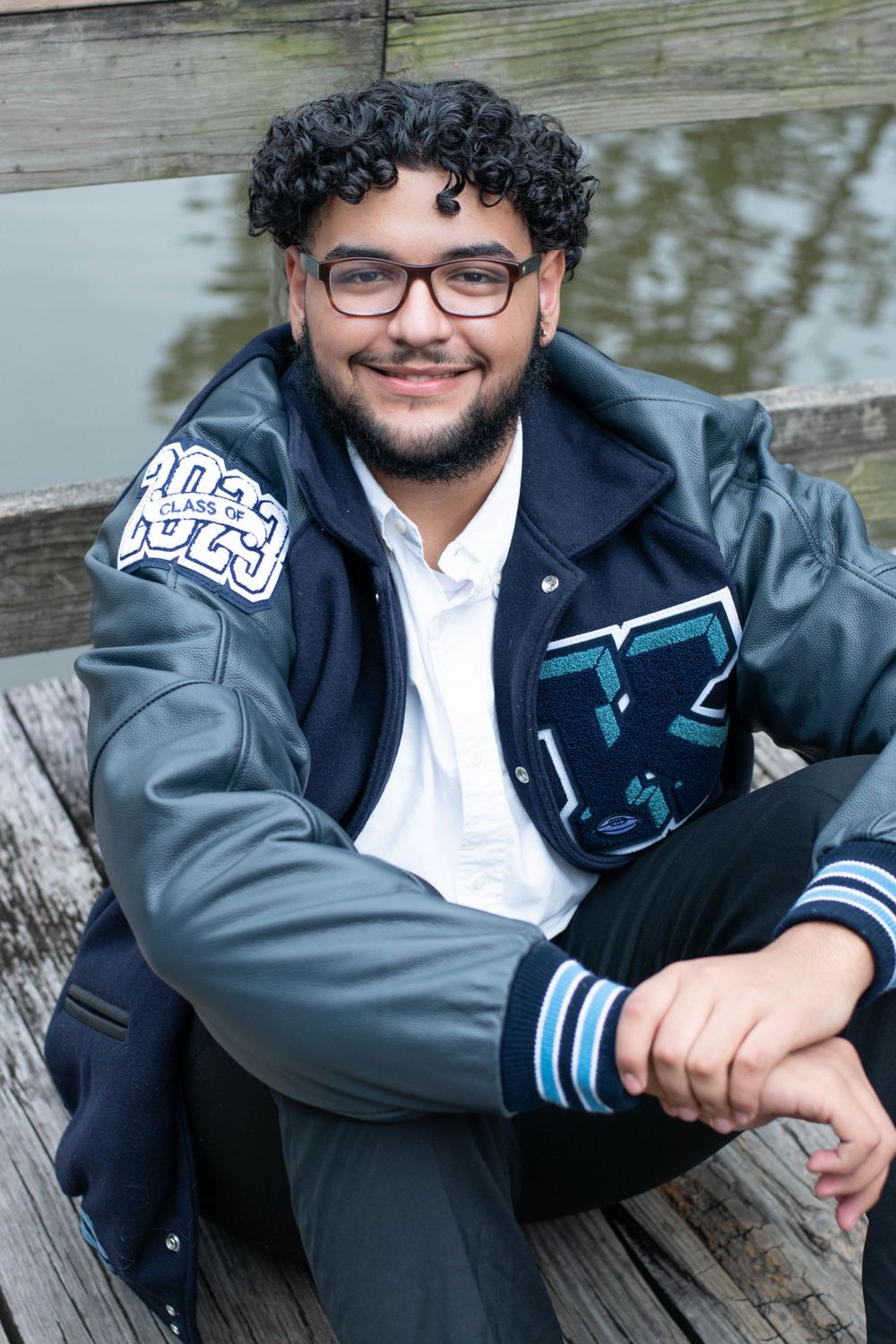 Graduate in varsity jacket sits casually on wooden dock by water.