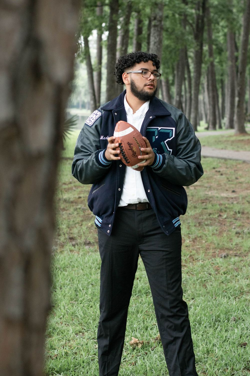Graduate in letterman jacket holds football while standing in wooded area.