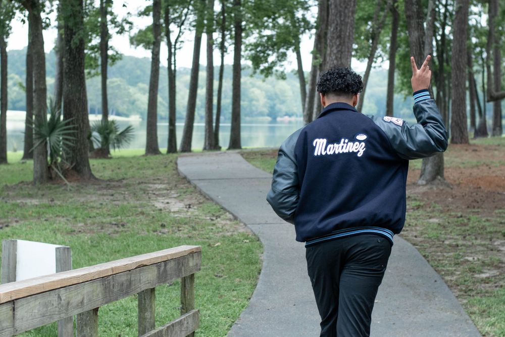 Graduate in a varsity jacket walks along a wooded path near a lake.