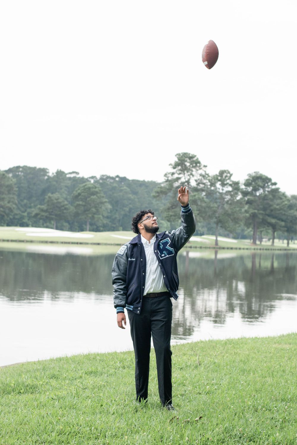 Graduate tosses football in the air by lakeside in graduation attire.