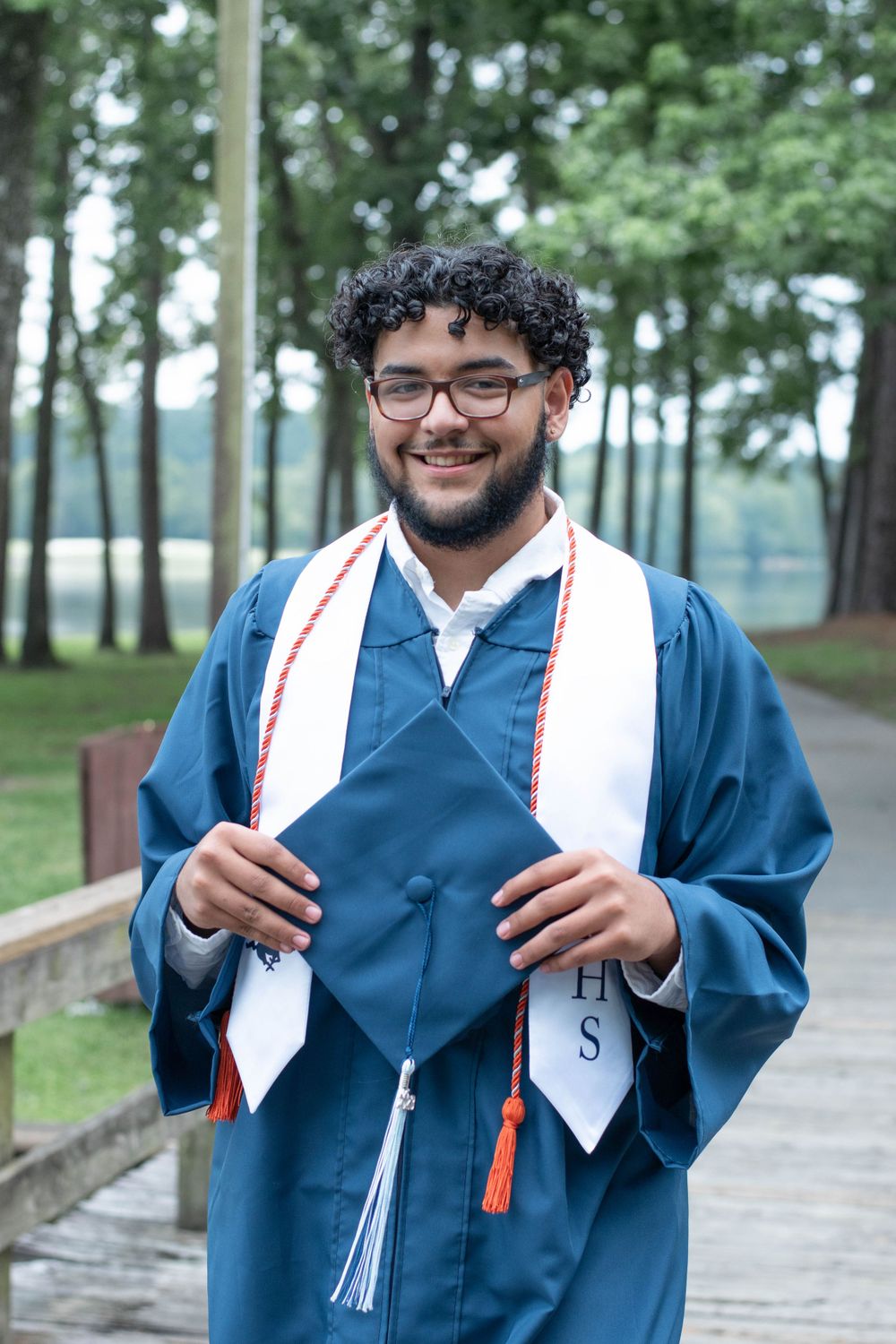Graduate in blue cap and gown with white stole smiles while posing outdoors.