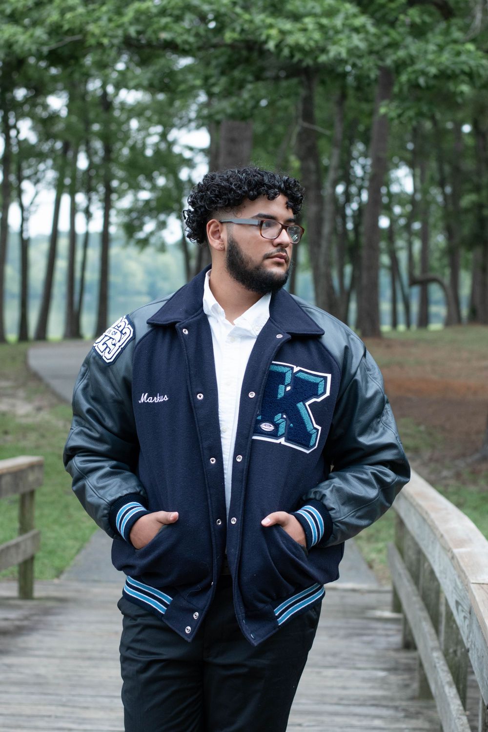 Graduate wearing letterman jacket poses on wooden boardwalk in wooded setting.
