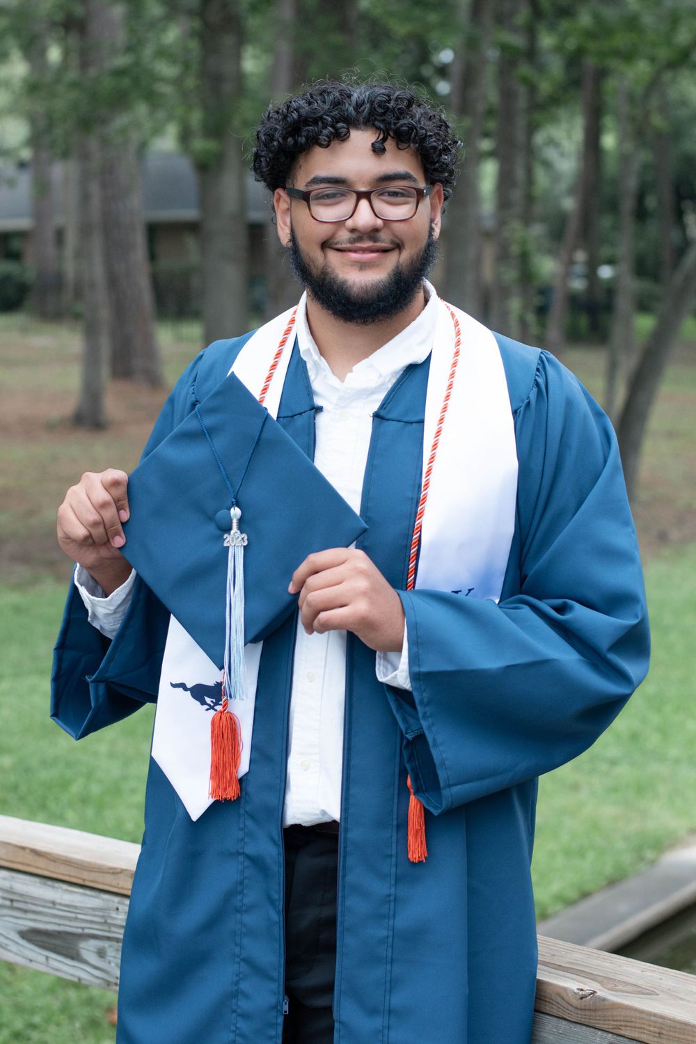 Graduate in blue gown displays cap and tassel while posing in outdoor setting.