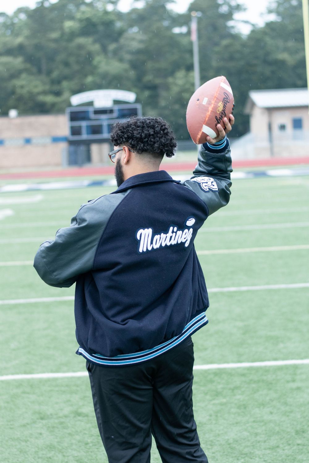 Graduate throws football while wearing varsity jacket on sports field.