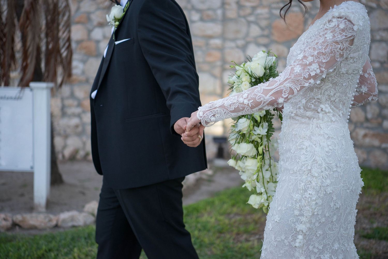 A detail shot of a bride and groom holding hands, showcasing her lace sleeve and his dark suit.