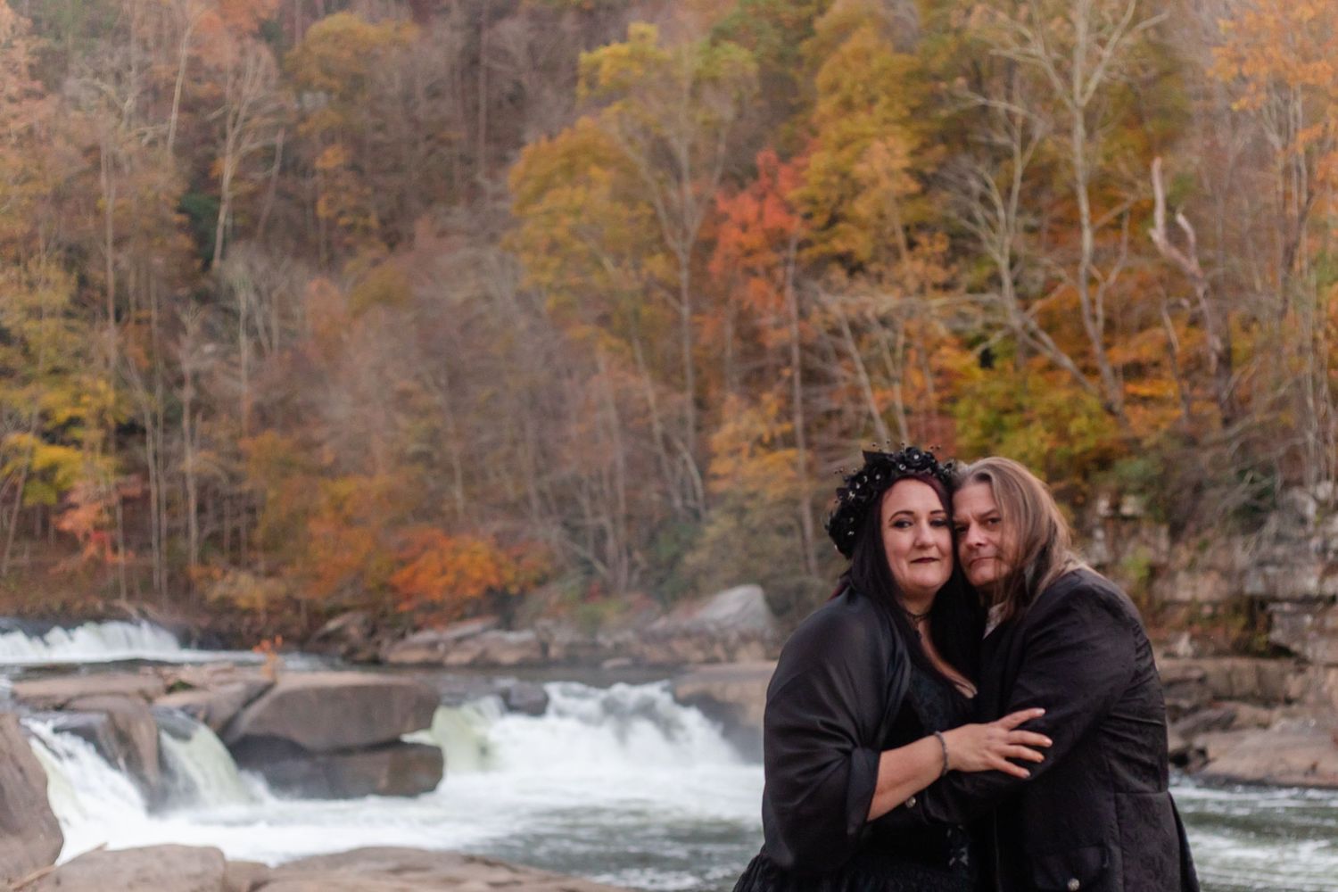 Two people embrace near a rushing waterfall surrounded by fall colors.