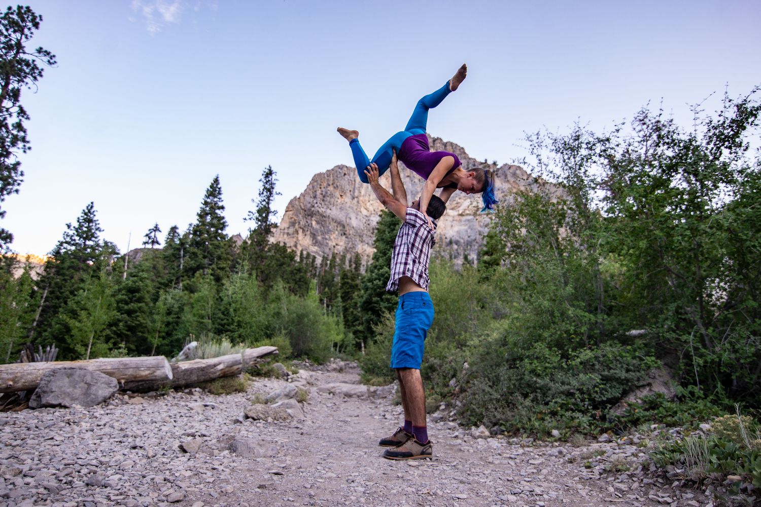 Acrobatic partner stunt with two people performing a balanced lift against a mountain backdrop.