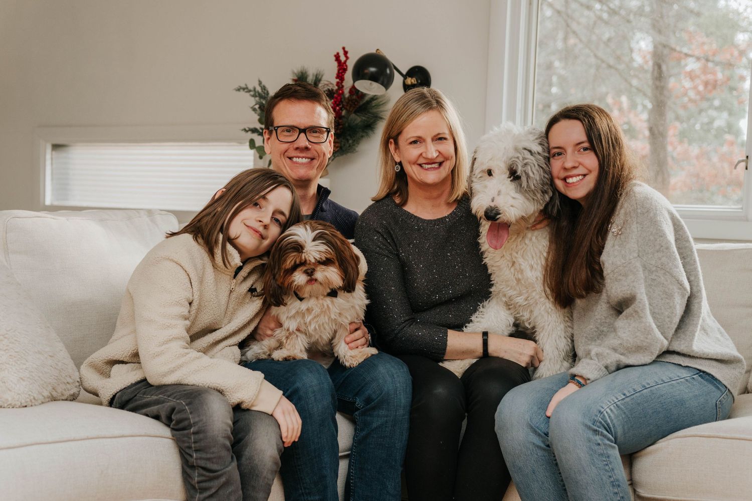 Family portrait on white couch with two fluffy dogs and holiday decorations.