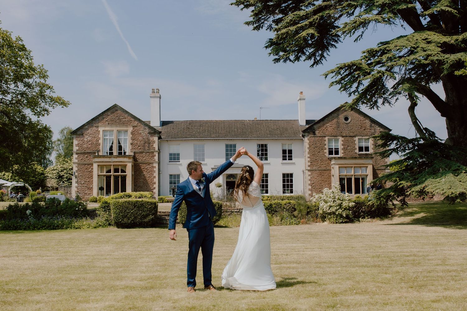 A couple celebrates on the lawn in front of a grand stone country manor house.