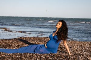 A woman in a blue dress relaxes on a rocky beach at sunset with the ocean in the background.