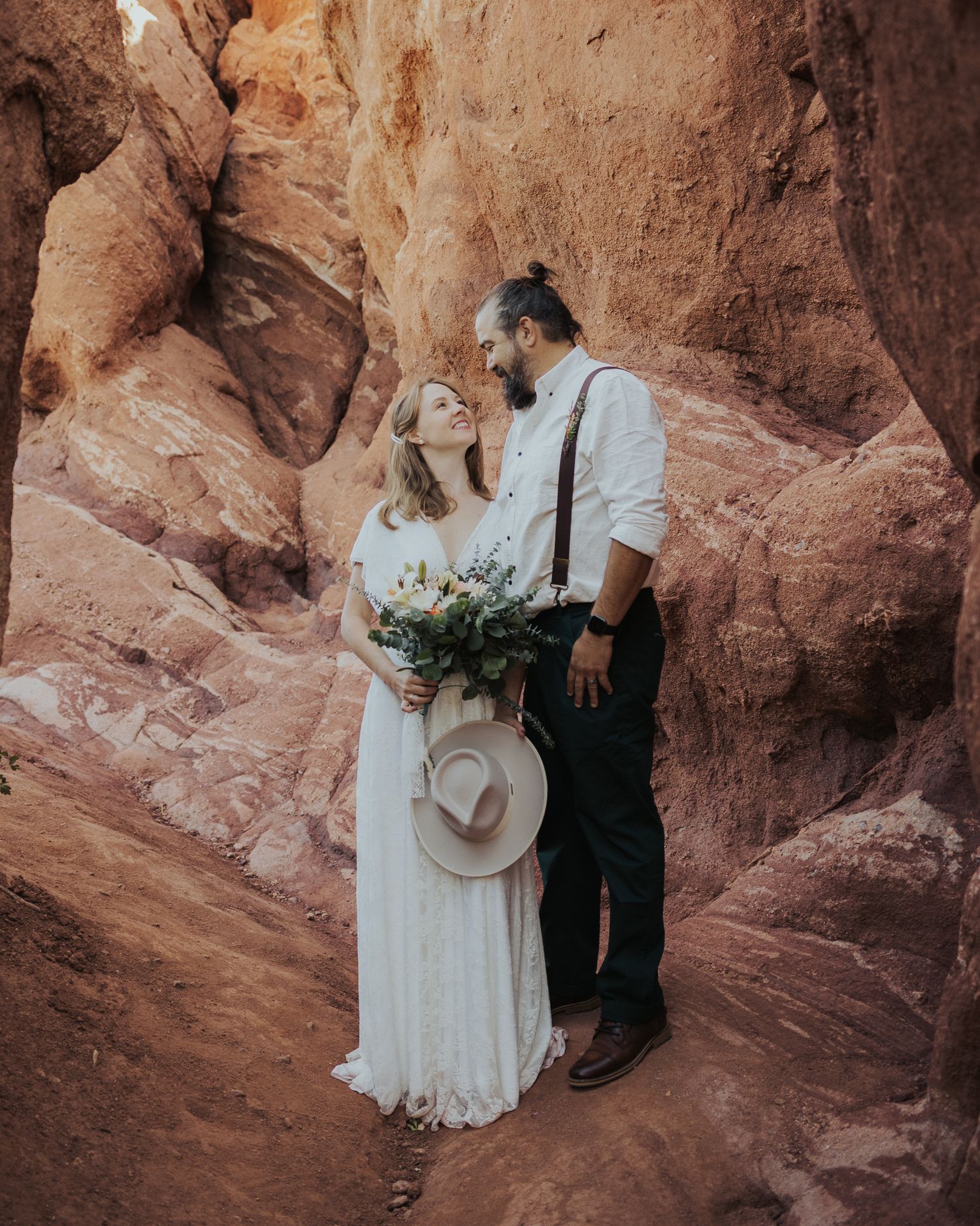 A couple in bohemian wedding attire poses in a red rock canyon holding a bouquet.