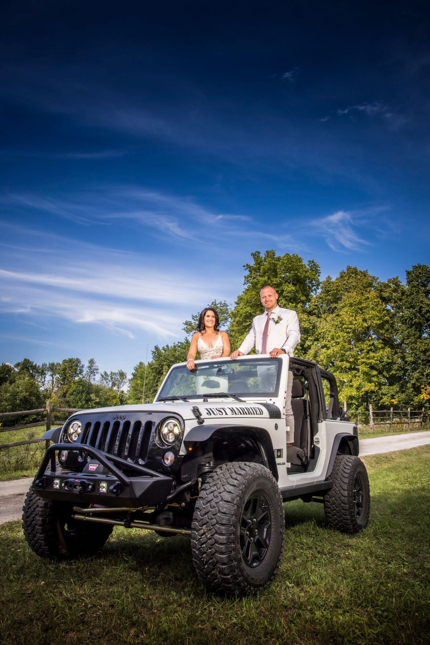 Wedding couple poses with white Jeep Wranglers on grassy hillside.