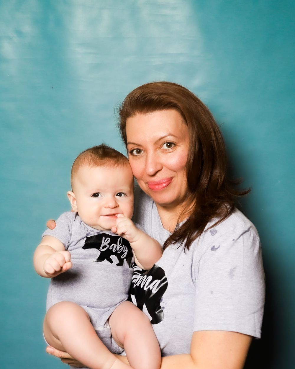 Portrait of mother and baby in matching grey outfits against teal background.