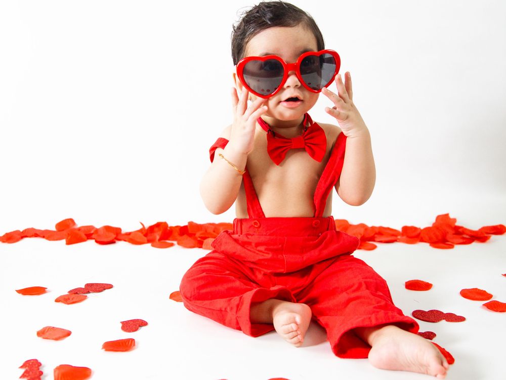 Baby wearing red suspenders and heart sunglasses surrounded by valentines decorations.