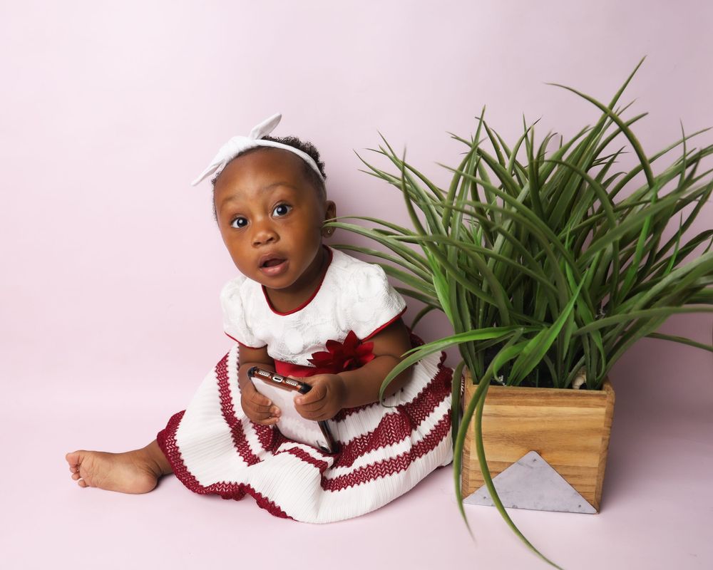 Baby in red and white dress sitting next to potted plant on pink background.